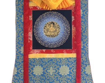 Puede incluir: Un tapiz vertical con un cuadrado central azul oscuro que presenta una ilustración de mandala circular dorada rodeada de escritura compleja. El textil está enmarcado por bordes de tela roja y amarilla y una base azul con motivos florales.