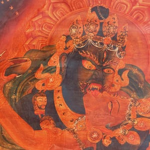 May include: A detailed thangka painting featuring a dark blue deity with a fierce expression, bared teeth, and a third eye, embracing a red figure. The artwork is rendered in vibrant shades of orange and red, adorned with intricate golden crowns.