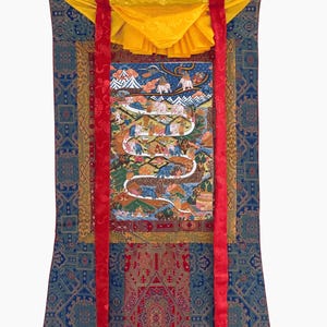 Può includere: Un arazzo thangka verticale con un dipinto centrale raffigurante un sentiero tortuoso con elefanti e figure, circondato da un bordo in tessuto multistrato blu, rosso e oro. Due lunghi nastri di seta rossa pendono da un baldacchino superiore giallo.