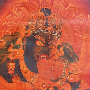 May include: A detailed painting featuring a dark-skinned deity with a fierce expression, bared teeth, and a third eye, embracing a lighter-skinned figure. The artwork is rendered in shades of red and orange, with a lotus-shaped halo background.
