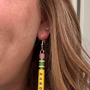 Peut inclure: Boucles d'oreilles fantaisie en forme de crayon. Les boucles d'oreilles sont jaunes avec des rayures vertes et noires, une gomme rose et le mot "NABORS" imprimé verticalement en noir. Elles sont suspendues à des crochets argentés.