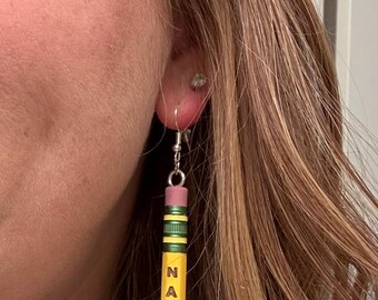 Puede incluir: Un par de pendientes con forma de lápiz. Los pendientes son amarillos con rayas verdes y negras, una goma de borrar rosa y la palabra "NABORS" impresa verticalmente en negro. Cuelgan de ganchos plateados.