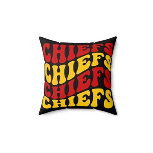 Pode incluir: Almofada quadrada preta com a palavra "CHIEFS" em fonte retrô vermelha e amarela repetida. Um item decorativo, provavelmente para um sofá ou cama.