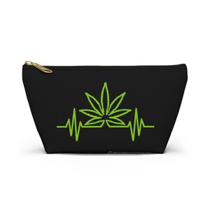 Könnte beinhalten: Schwarze Reißverschlusstasche mit einem grünen Cannabisblatt-Design, das in eine Herzschlag-Grafik integriert ist. Die Tasche hat einen goldfarbenen Reißverschluss und eine rechteckige Form.