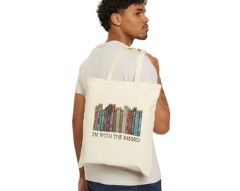 Puede incluir: Bolso tote de color crema con un gráfico de una fila de libros de varios colores. El texto "I'M WITH THE BANNED" está impreso debajo de los libros. El bolso tiene correas largas para llevar al hombro.