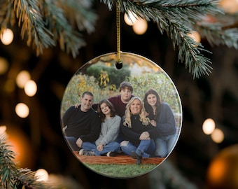 Peut inclure: Ornement de Noël rond en céramique avec un portrait de famille. L'ornement est suspendu à une ficelle dorée sur fond de sapin de Noël avec des lumières floues. La famille est assise sur une couverture.