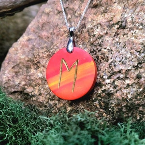 Peut inclure: Un pendentif rond rouge et orange avec un 'M' doré peint dessus. Le pendentif est suspendu à une chaîne argentée.