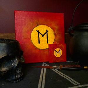 May include: Two canvas paintings with a red background and a yellow circle containing a black rune symbol. The rune symbol is the letter "M".