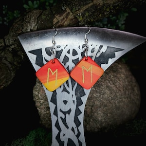 Peut inclure: Une paire de boucles d'oreilles carrées rouges, orange et jaunes avec une rune M dorée peinte sur chacune. Les boucles d'oreilles sont suspendues à des crochets argentés.