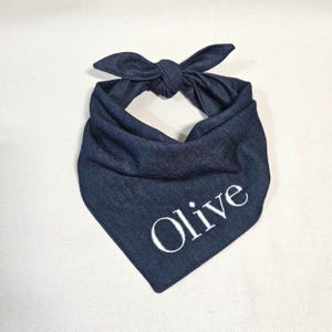 Op de afbeelding: Een donkerblauwe denim bandana voor huisdieren in een driehoekige vorm met een knoop aan de bovenkant. De stof heeft een zichtbare geweven textuur en de naam Olive is geborduurd in witte hoofdletters en kleine letters op het onderste deel.