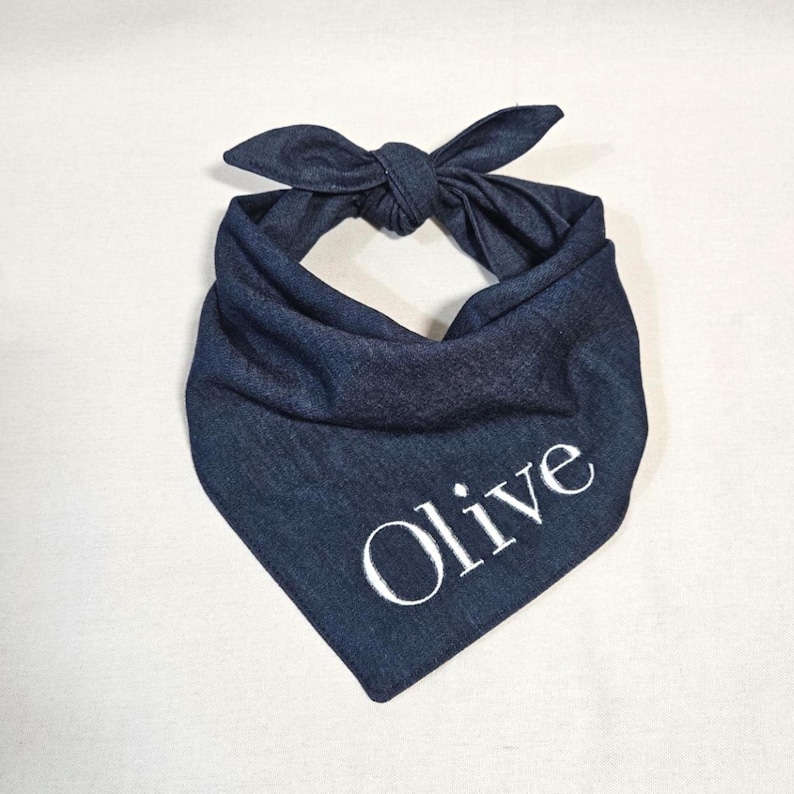 Op de afbeelding: Een donkerblauwe denim bandana voor huisdieren in een driehoekige vorm met een knoop aan de bovenkant. De stof heeft een zichtbare geweven textuur en de naam Olive is geborduurd in witte hoofdletters en kleine letters op het onderste deel.