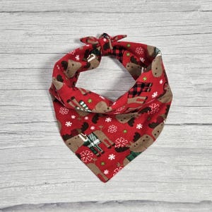 May include: Red bandana with a Christmas moose print. The bandana is tied in a knot at the top and has a brown trim.