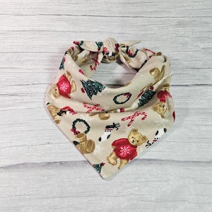 May include: A beige bandana with a Christmas-themed print featuring teddy bears, Christmas trees, candy canes, and wreaths. The bandana is tied in a knot at the top.