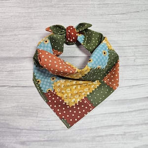 May include: A patchwork dog bandana with a bow. The bandana features a variety of fabrics in shades of green, orange, yellow, and blue. The fabrics have floral and polka dot patterns. The bow is made of green fabric with a red center.