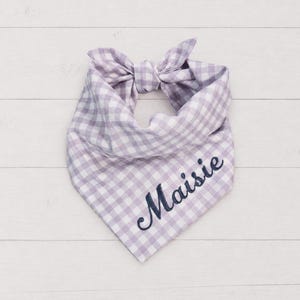 May include: A light purple and white gingham patterned pet bandana featuring a tied knot at the top. The fabric is embroidered with the name Maisie in a dark navy blue cursive script font, positioned diagonally across the lower right section.