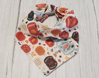 Peut inclure: Un bandana blanc avec un imprimé sur le thème de l'automne avec des citrouilles orange, rouge et bleues, des feuilles et des étoiles. Le bandana est noué en haut.