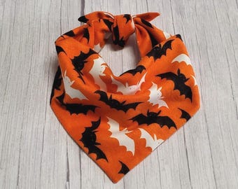 May include: A triangular pet bandana featuring a vibrant orange background patterned with repeating black and white bat silhouettes. The fabric is tied at the top into a knot, creating a folded shape with pointed edges and a finished hem.