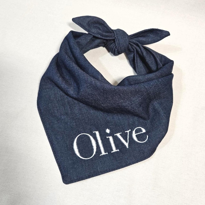 Op de afbeelding: Een donkerblauwe denim bandana voor huisdieren in een driehoekige vorm met een knoop aan de bovenkant. De stof heeft een textuur en is voorzien van de naam Olive, geborduurd in witte hoofdletters en kleine letters in het midden.