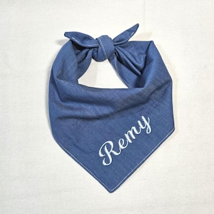 Op de afbeelding: Een blauwe spijkerstof bandana voor huisdieren in een driehoekige vorm met een knoop aan de bovenkant. De stof heeft zichtbare witte stiksels langs de randen en de naam Remy is in wit cursief schrift op het onderste deel geborduurd.
