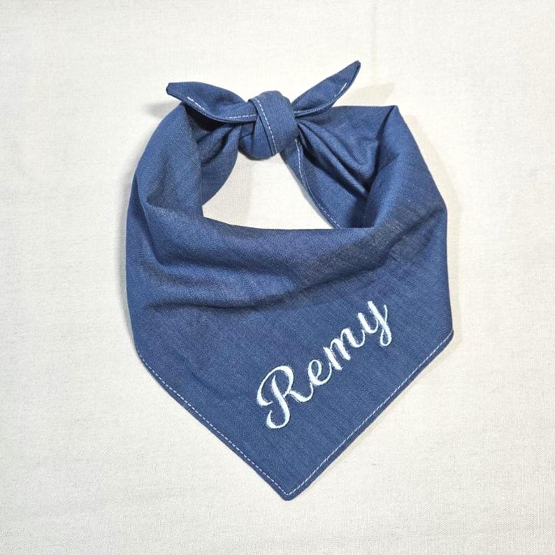 Op de afbeelding: Een blauwe spijkerstof bandana voor huisdieren in een driehoekige vorm met een knoop aan de bovenkant. De stof heeft zichtbare witte stiksels langs de randen en de naam Remy is in wit cursief schrift op het onderste deel geborduurd.