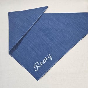 Op de afbeelding: Een driehoekige blauwe spijkerstof bandana met de naam Remy geborduurd in wit cursief schrift. De stof heeft een zichtbare geweven textuur en wit gestikte randen, gevouwen om de gepersonaliseerde belettering duidelijk op de voorzijde te tonen.