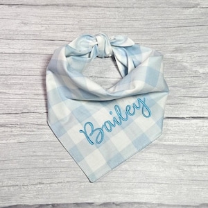 May include: A light blue and white gingham dog bandana with the name "Bailey" embroidered in a matching light blue thread. The bandana is tied at the top, creating a bow. The fabric has a classic plaid pattern.
