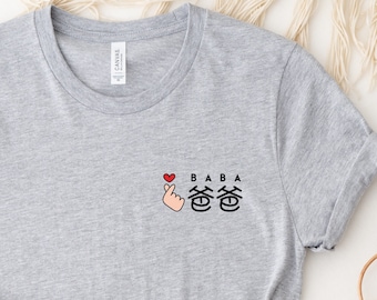 Puede incluir: Camiseta gris con un pequeño gráfico de una mano haciendo un gesto de corazón con el texto "BABA" y dos caracteres chinos.