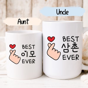 May include: Two white ceramic mugs with black text and a pink hand gesture with a red heart. The mug on the left says "Best Eomo Ever" and the mug on the right says "Best Sangchun Ever".