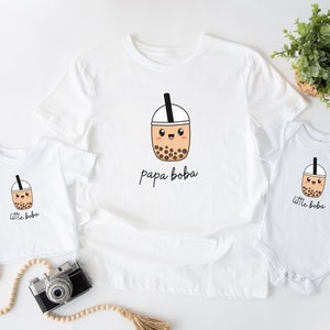 May include: White t-shirts with a cartoon boba tea design. The adult shirt says "papa boba" and the child shirt says "little boba".