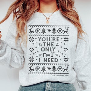 May include: A light grey sweatshirt featuring a festive pattern with reindeer, snowflakes, hearts, and Christmas trees. The text reads "YOU'RE THE ONLY n=1 I NEED" with "99% confidence level" below. A comfortable, holiday-themed top.