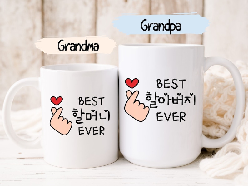 May include: Two white ceramic mugs with black text and a red heart and hand gesture. The text on the mugs reads "Best 할머니 Ever" and "Best 할아버지 Ever".