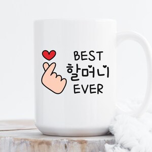 May include: White ceramic mug with a black and white design that says "Best  하얘니  Ever" with a red heart and a pink hand making a heart sign.