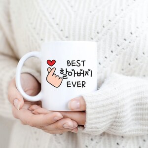 May include: White ceramic mug with a black and red design. The mug features a hand gesture with a heart and the text "BEST 할아버지 EVER".