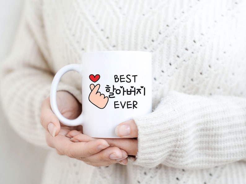 May include: White ceramic mug with a black and red design. The mug features a hand gesture with a heart and the text "BEST 할아버지 EVER".