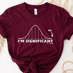 Puede incluir: Camiseta burdeos con un gr&aacute;fico de curva de campana blanca y el texto "I'M SIGNIFICANT p < 0.01". La palabra "ME" est&aacute; al lado de una flecha que apunta a la curva de campana. La camiseta es de material suave.