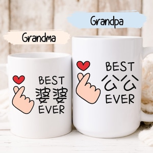 Peut inclure: Deux mugs en céramique blanche avec du texte et des illustrations noirs. Un mug dit "Grand-mère" et "Meilleur de tous les temps" en anglais et en caractères chinois. L'autre mug dit "Grand-père" et "Meilleur de tous les temps". Les deux ont un cœur rouge et un geste de la main.