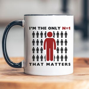 May include: White ceramic mug with a black rim and handle. The mug features a graphic of a red stick figure standing out from a crowd of black stick figures. The text reads "I'm the only N=1 that matters".