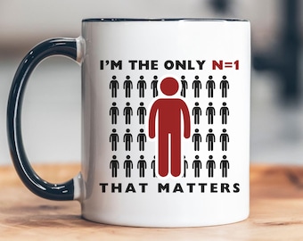 May include: White ceramic mug with a black rim and handle. The mug has a graphic of a red stick figure standing out from a crowd of black stick figures. The text reads "I'm the only N=1 that matters".