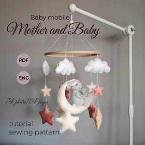 Op de afbeelding: Een witte babymobiel met een houten ring en wolken, sterren en een halve maan van vilt. De mobiel is gelabeld "Babymobiel Moeder en Baby" en bevat een PDF-naai-patroon met 74 foto's en 24 pagina's.