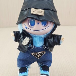May include: A blue plush toy creature dressed in a black faux-leather jacket, denim dungarees, and a matching hat. The outfit features a "PPAD" logo on the dungarees and a "Fashion Handmade" label on the hat. The toy is wearing blue and white trainers.