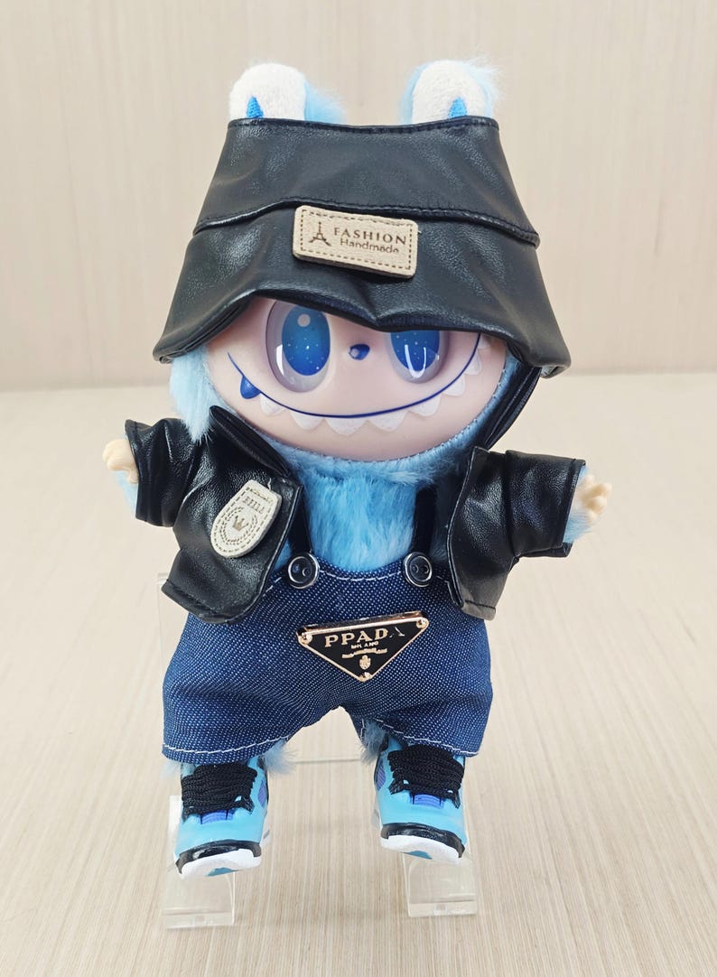 May include: A blue plush toy creature dressed in a black faux-leather jacket, denim dungarees, and a matching hat. The outfit features a "PPAD" logo on the dungarees and a "Fashion Handmade" label on the hat. The toy is wearing blue and white trainers.
