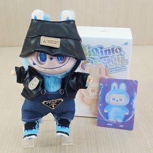 May include: A blue plush toy dressed in a black leather jacket, denim dungarees, and blue trainers. The toy has a black hat and is positioned in front of a box with the words "Biginto Energy" and a card that says "HOPE".