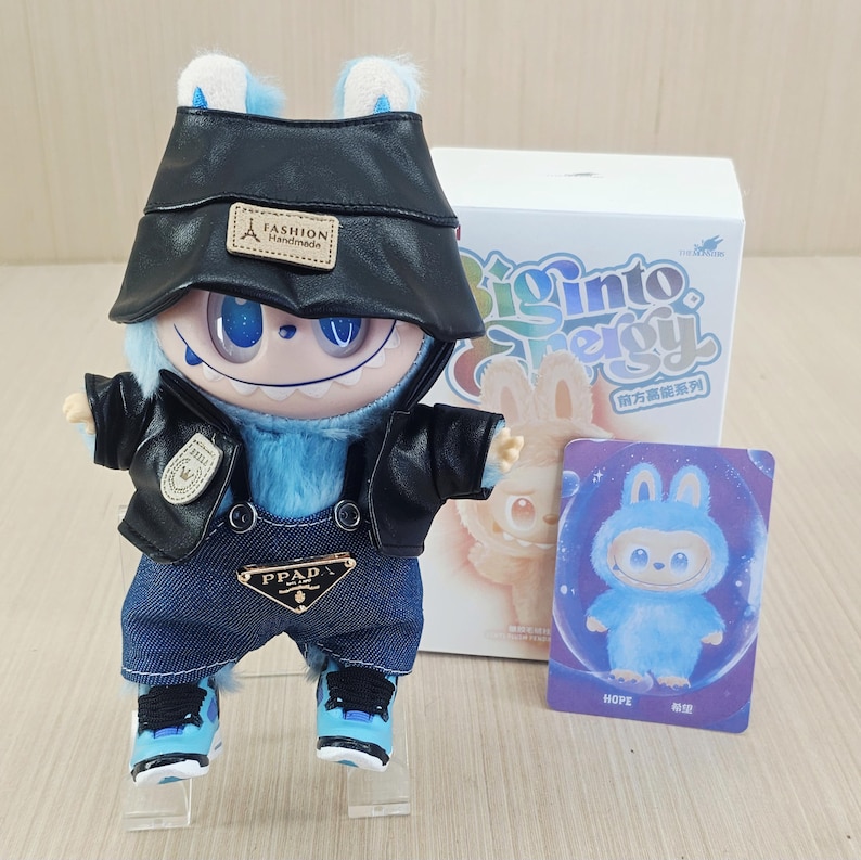 May include: A blue plush toy dressed in a black leather jacket, denim dungarees, and blue trainers. The toy has a black hat and is positioned in front of a box with the words "Biginto Energy" and a card that says "HOPE".