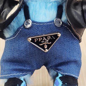 May include: A plush toy figure with light blue fur, wearing a denim overall with the "PPADA" logo, a black leather jacket, and blue trainers with black laces. The figure has a white-toothed grin.