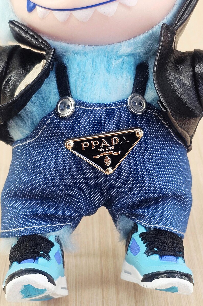 May include: A plush toy figure with light blue fur, wearing a denim overall with the "PPADA" logo, a black leather jacket, and blue trainers with black laces. The figure has a white-toothed grin.