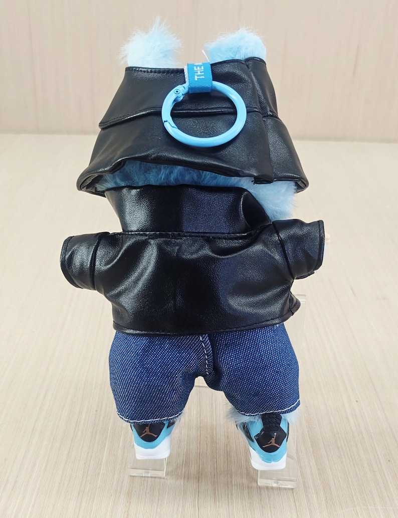 May include: A plush toy wearing a black faux leather jacket, denim shorts, and trainers. The toy has a blue fur head with a matching hat and a blue ring clip. The trainers have black and blue accents.