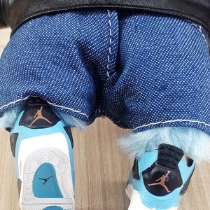 May include: A doll wearing blue denim jeans, a black leather jacket, and light blue and white trainers. The trainers have a black and orange logo and a light blue sole.