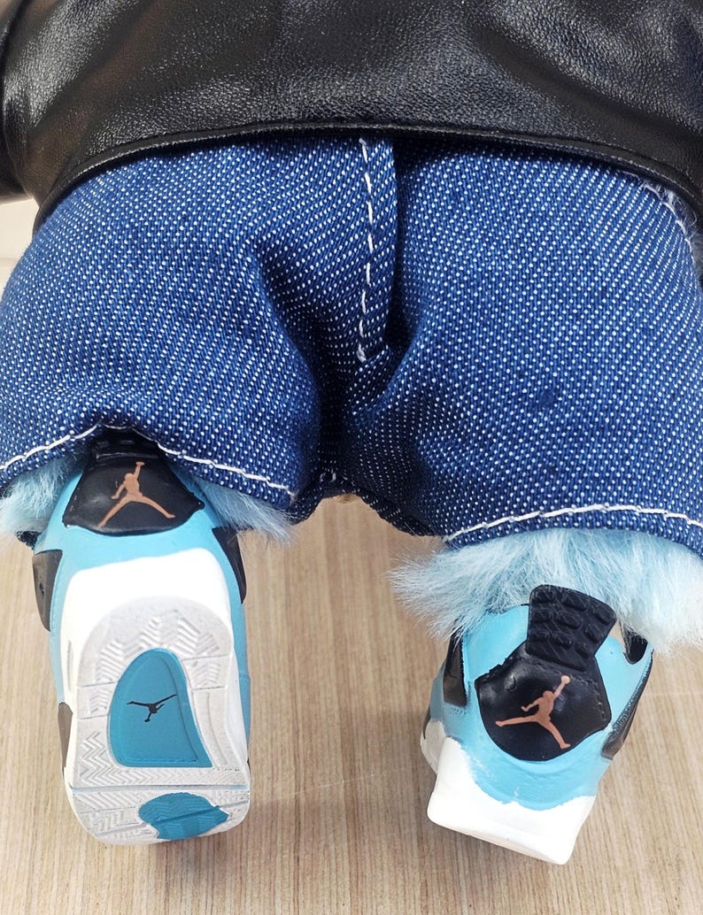 May include: A doll wearing blue denim jeans, a black leather jacket, and light blue and white trainers. The trainers have a black and orange logo and a light blue sole.