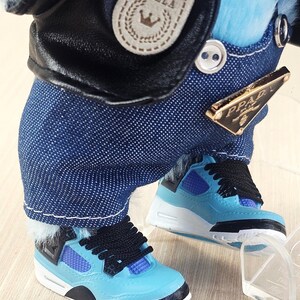 May include: A doll dressed in a black leather jacket with a "BELLA" patch and blue denim overalls. The doll is wearing light blue trainers with black laces and accents. A gold-coloured "PRADA" tag is visible on the overalls.