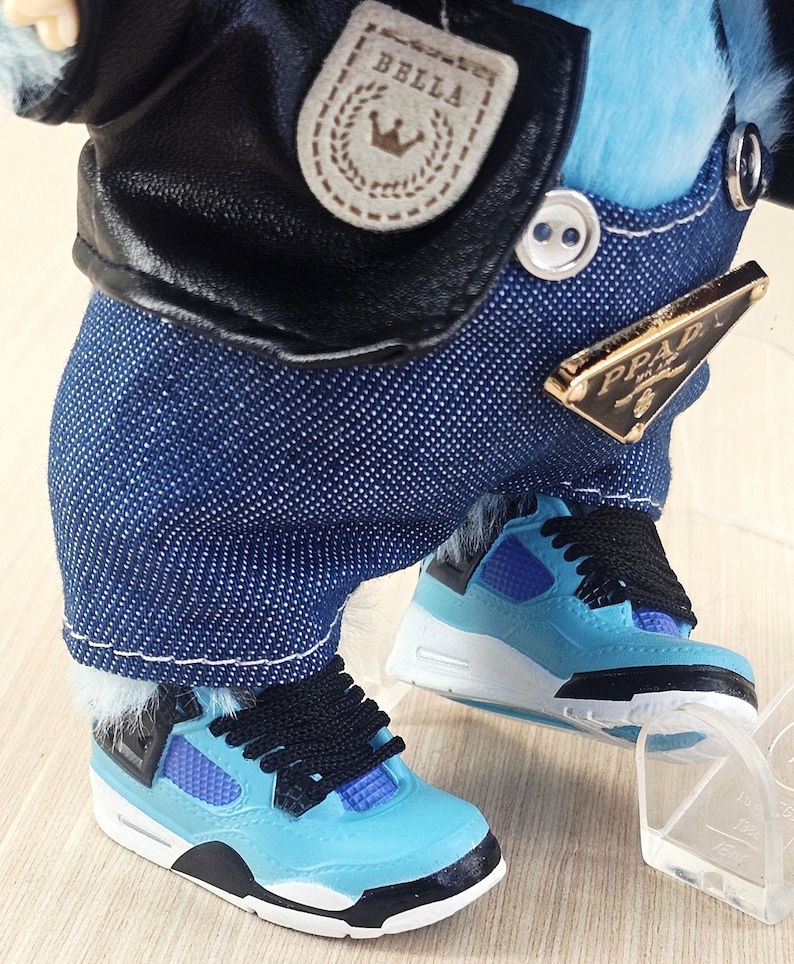 May include: A doll dressed in a black leather jacket with a "BELLA" patch and blue denim overalls. The doll is wearing light blue trainers with black laces and accents. A gold-coloured "PRADA" tag is visible on the overalls.